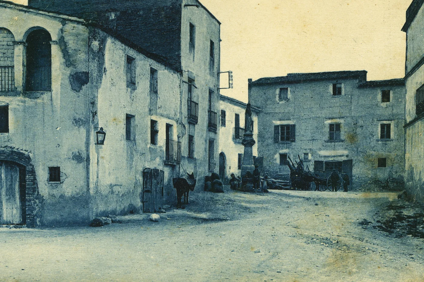 Postal antiga de la plaça de les Moreres de Torrelles de Foix, amb cases de pedra, una mula i carros al fons
